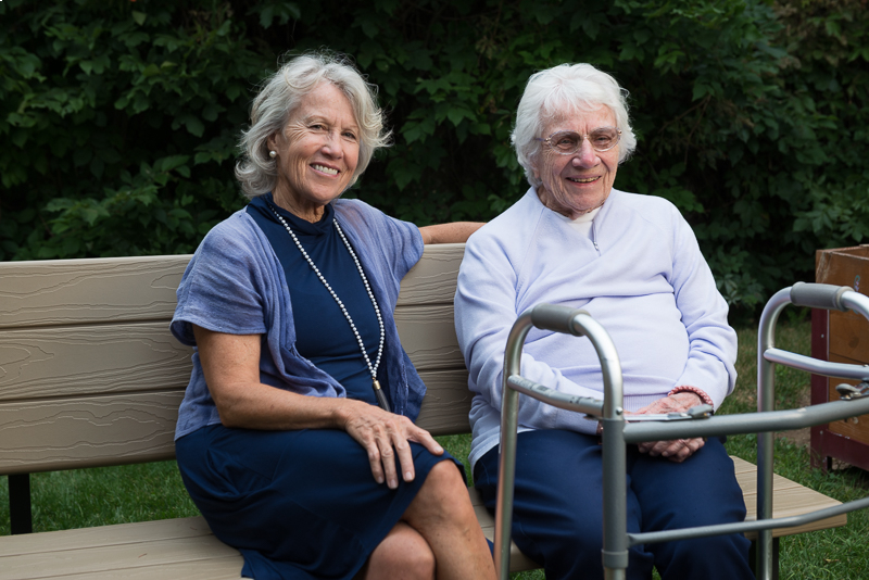 Two smiling older women sitting on a park bench next to a mobility walker.